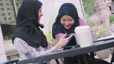 Two young asian Muslim females wear hijab sitting at the cafe, using smartphone , buying new gadget, friend meet up appointment, modern technology, posting photos online, friend reunion happiness