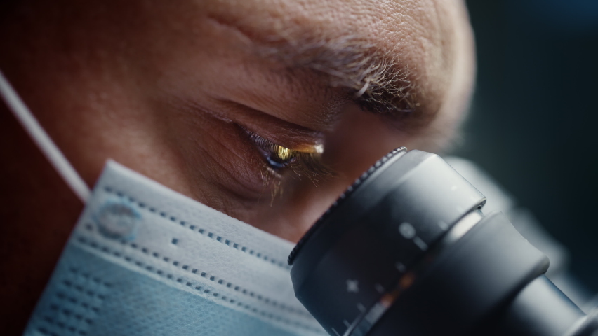 Ultra Macro Close Up Footage of a Male Scientist Wearing Medical Mask and Looking into the Microscope. Microbiologist Working on Molecule Samples in Modern Laboratory with Technological Equipment.