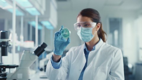Beautiful Female Scientist Wearing Face Mask and Glasses Looking a Petri Dish with Genetically Modified Sample Chemicals. Microbiologist Working in Modern Laboratory with Technological Equipment.