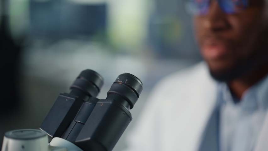 Macro Close Up Footage of a Handsome Black Male Scientist Wearing Glasses and Looking into the Microscope. Microbiologist Working on Molecule Samples in Modern Laboratory with Technological Equipment. Royalty-Free Stock Footage #1065058000