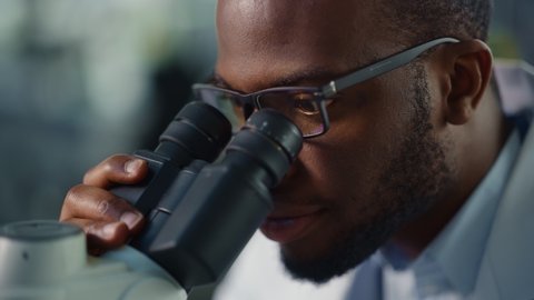Macro Close Up Footage of a Handsome Black Male Scientist Wearing Glasses and Looking into the Microscope. Microbiologist Working on Molecule Samples in Modern Laboratory with Technological Equipment.
