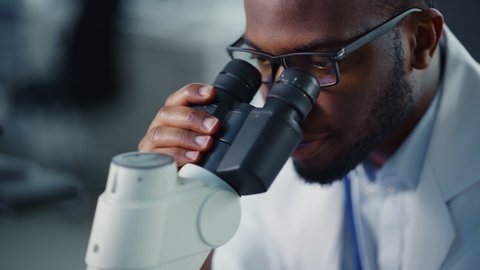 Close Up Footage of a Black Male Scientist Putting a Petri Dish with Genetically Modified Sample Chemicals Under a Microscope. Microbiologist Working in Modern Laboratory with Technological Equipment.