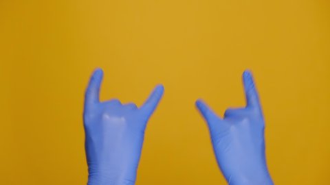 Rock-n-roll and pandemic concept. Close up male hands in medical protective blue gloves waving with horns gesture, isolated on yellow studio background with copy space for advertisement. Body language