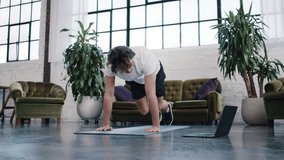 Young sporty man does a home workout on a yoga mat while watching a fitness video on a laptop in a stylish, modern apartment. Sports from home.
