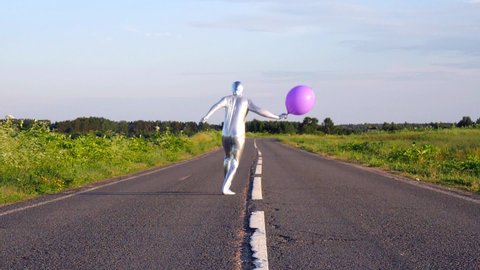 Tall slender man in silver suit is fully dressed in tight silver suit, runs slowly down  middle of an empty road, with pink, purple large inflatable ball in his hands. Freak, an alien, unusual, UFO.