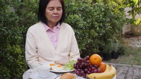 Happy senior Asian woman sitting in the backyard garden, going to have breakfast, using smartphone, laughing, waving hand, making video call, talking, and smiling. Outdoors, happy retirement concept