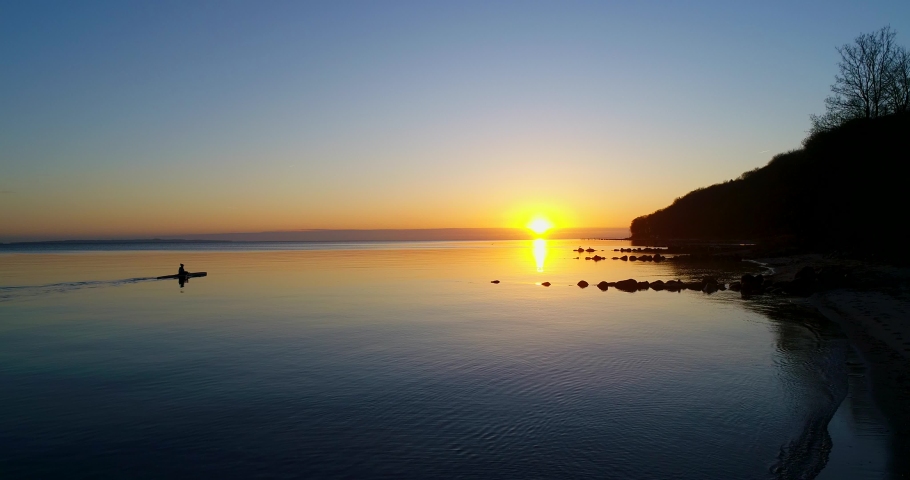 Lonely canoeing adventure. Silhouet of a person canoeing early in the morning along the calm shore of Århus Denmark towards the sunrise.  Peaceful scenery. Paddling along the Danish coast, Europe.