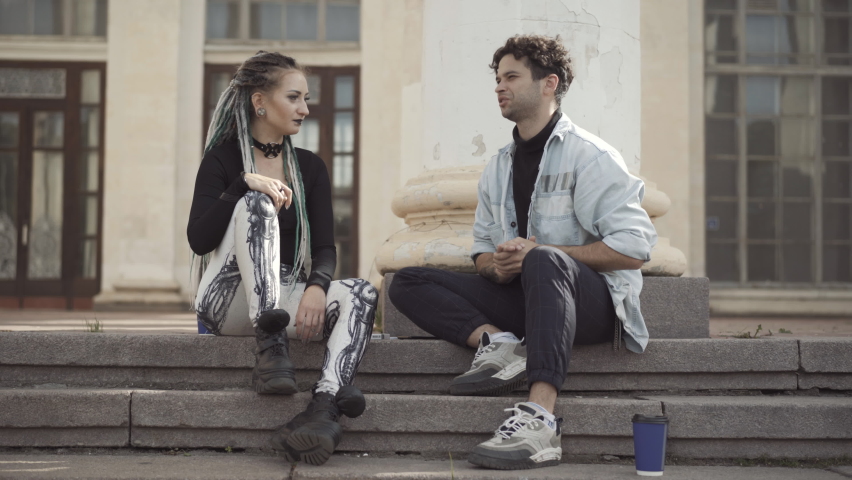 Excited young Caucasian hippie sitting on urban stairs with goth woman and talking. Portrait of relaxed millennial couple dating outdoors on sunny summer day. Relationship and subculture concept.