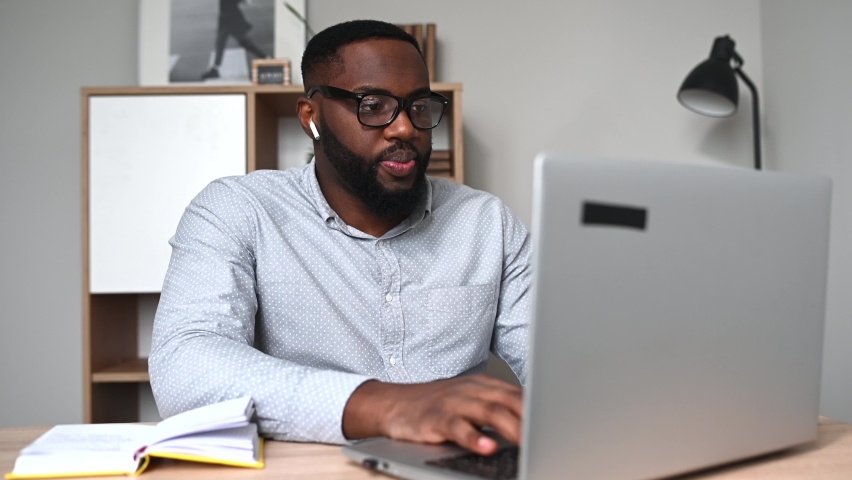 Cheerful young multiethnic guy in glasses using laptop sitting at the desk in home office. An African entrepreneur websurfing, answering email, chatting online with a smile Royalty-Free Stock Footage #1065081589