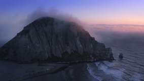 Scenic rock on the ocean coast covered by the pink and purple clouds moving slowly in the sky and golden sun shining through the clouds with at sunset. Flying in heavenly beautiful sunny cloudscape 4K