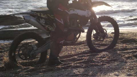 Biker wearing protective equipment slips in golden quicksand on beach against waving sea and bright sunlight closeup slow motion