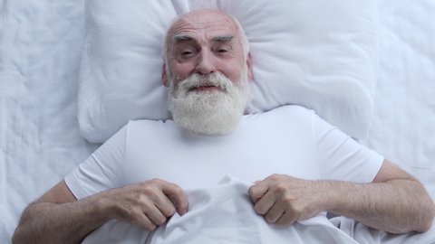 Cheerful greyhaired man lying in bed, feeling happy, thinking of funny moments