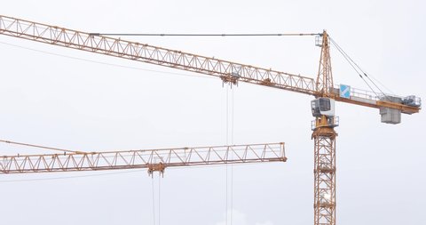Two yellow tower crane jibs with working trolley on a construction site against white cloudy sky 