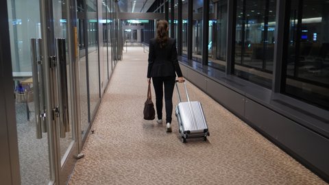 Tired passenger woman walk at empty terminal passage with small trolley case, camera follow behind. Modern airport arrival area, glass wall on right side, dark hour, late flight arrivals