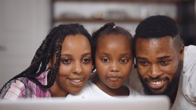 Happy afro-american family surfing internet together on laptop at home. African mother, father and little daughter shopping online or watching movie on computer