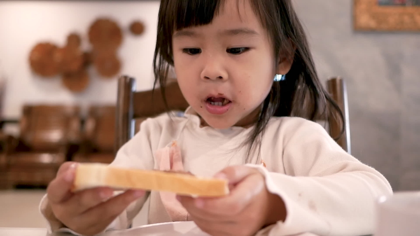 Cute little girl eating tasty bread with jam in kitchen in the morning.
