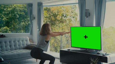 Woman infront of green screen TV mock up doing squad workout fitness in modern living room