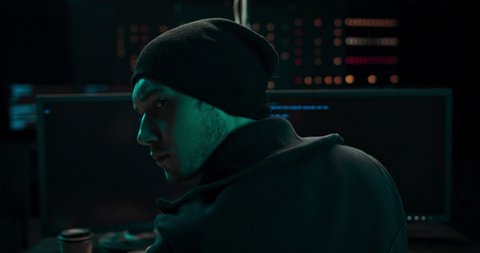 Shot from the Back to Hooded Hacker Breaking into Corporate Data Servers from His Underground Hideout. Wanted anonymous programmer coding virus ransomware using computers. Multiple Displays Background