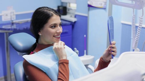 Female patient in the dental clinic is satisfied with the results of the procedures, looking at mirror and smiling. Visiting dentist. Healthcare and medicine concept.