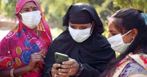 Close-up of group of women in ethnic clothes, Muslim Hijab and Hindu Sari with face mask discuss share using smart phone mobile technology communication device during coronavirus covid-19 lockdown