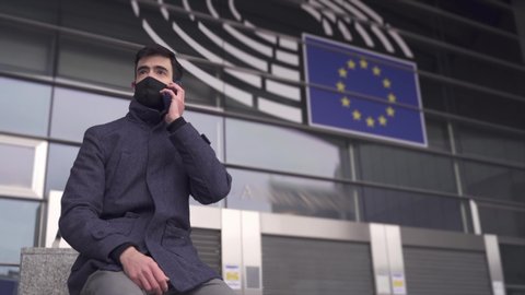 Young man with face mask in front of European Parliament in Brussels on a phone call. EU Parliamentary and Businessman in times of Coronavirus Crisis in Europe