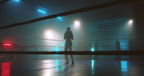 Female active sporty boxer training at ring in darkness and boxing alone in air. Young girl kickboxer practising attack moves. Woman in gloves punching like fighting. Fighter working out.