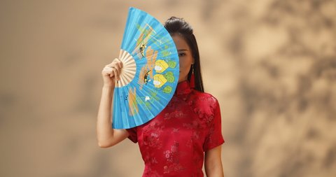 Portrait shot of young Asian beautiful and cheerful woman in red traditional clothes cheongsam clothing her face with blue fan and opening as smiling happily. Geisha style concept.