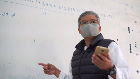 Professor or teacher female Asian with mask prevent COVID-19, Teacher teaching or speaking using smartphone for e-classroom or e-learning explaining at white board in classroom university in education