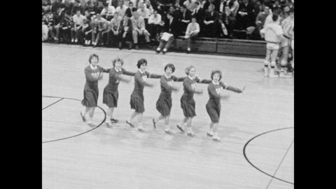 1960s: Cheerleaders at basketball game. Basketball players on bench. Cheerleaders perform for whole gymnasium.