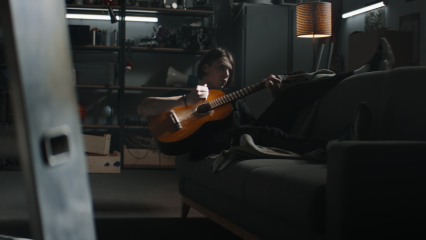 MED HANDHELD Caucasian teenager boy lying on a sofa in his home garage hideout, playing guitar. Shot with 2x anamorphic lens