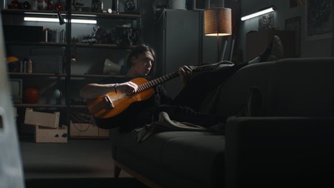 MED HANDHELD Caucasian teenager boy lying on a sofa in his home garage hideout, playing guitar. Shot with 2x anamorphic lens