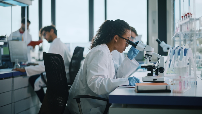 Medical Science Laboratory: Beautiful Black Scientist Looking Under Microscope Does Analysis of Test Sample. Diverse Team of Young Specialists, Using Advanced Technology Equipment. Side View Zoom Royalty-Free Stock Footage #1065136591