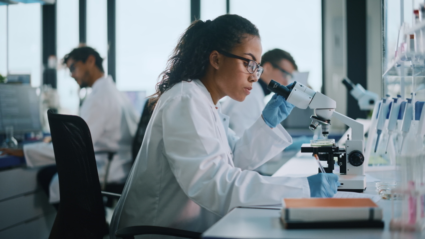 Medical Science Laboratory: Beautiful Black Scientist Looking Under Microscope Does Analysis of Test Sample. Diverse Team of Young Specialists, Using Advanced Technology Equipment. Side View Zoom | Shutterstock HD Video #1065136591