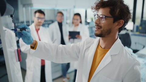 Medical Science Laboratory: Handsome Latin Male Scientist Writes Detailed Project Data Analysis on the Board, His Diverse Team of Colleague Listens. Young Scientists Solving Problems. Elevated Shot