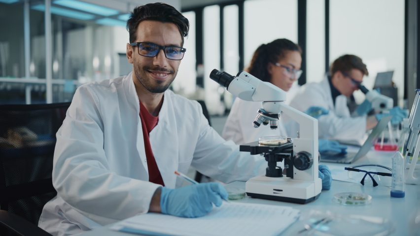 Medical Science Laboratory: Handsome Latin Scientist Looking Under Microscope, Looking at Camera and Smiling Charmingly. Young Biotechnology Specialist, Using Advanced Equipment. Slow Motion Zoom in | Shutterstock HD Video #1065136720