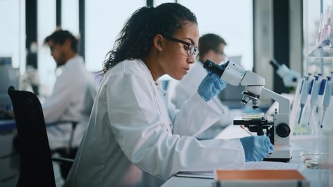Medical Science Laboratory: Beautiful Black Scientist Looking Under Microscope, writes down Analysis Data. Young Biotechnology Specialist, Using Advanced Equipment. Side View Slow Motion Zoom in