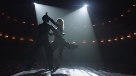 Attractive couple of ballroom dancers dancing passionate latin american dance cha cha cha. Silhouettes of two dancers on a smoky Stage with spot light. Anamorfic lens