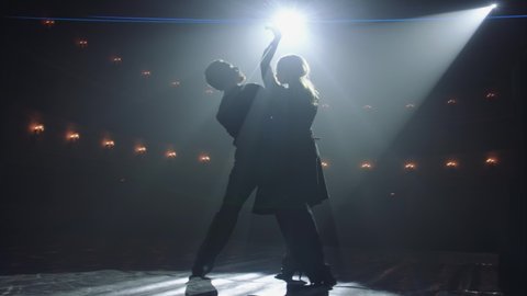 Attractive couple of ballroom dancers dancing passionate latin american dance cha cha cha. Silhouettes of two dancers on a smoky Stage with spot light. Anamorfic lens