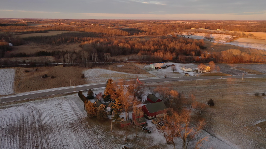 Aerial view of farm house in Wisconsin in spring winter season, agricultural field covered in snow, American flag waves in the front yard. Establishing shot of Midwestern countryside