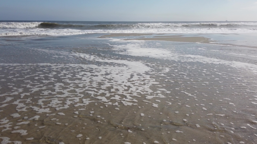 Ocean water retreats back to the sea after washing up on the sandy beach. Rough ocean waves are seen in the background