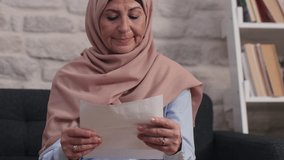 Sad middle-aged woman in turban is reading a letter at her home. The old woman with a headscarf cries at the letter she read and receives sad news.Close up, slow motion video.