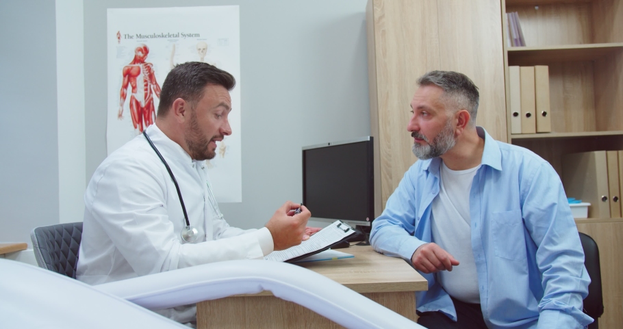 Side view. Portrait of Caucasian focused male doctor speaking and consulting middle-aged man patient about illness or surgery sitting in hospital cabinet. Handsome physician giving advice on health | Shutterstock HD Video #1065150571