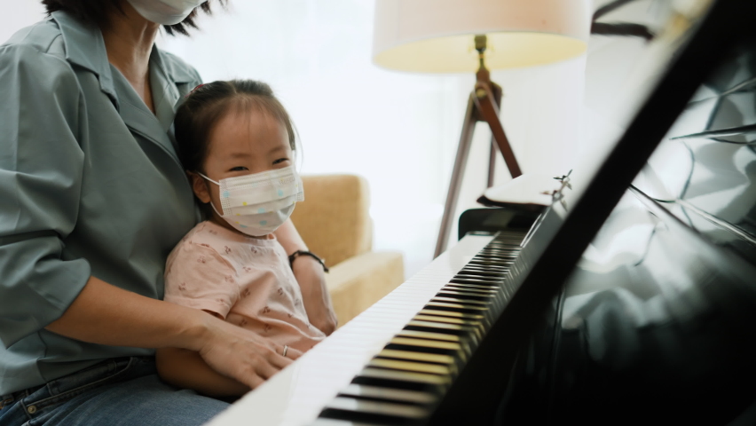 Medium close up shot of Asian young woman and toddler 3 years old girl wearing face mask look to camera and show finger sign of victory after playing piano, New normal with face mask after covid-19. Royalty-Free Stock Footage #1065160879