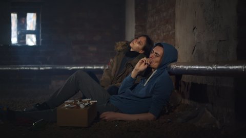 Drug addicted man and woman smoking sitting on floor in dirty basement. People resting after using heavy drugs. Trafficking, crime, addiction concept