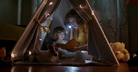Happy caucasian mom, cute baby boy and funny dog spending time together at home, reading a book, using a flashlight in a cozy tent - happy family 4k footage