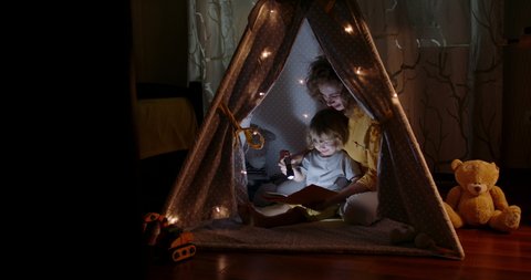 Happy caucasian mom and her cute baby boy spending time together at home, reading a book, using a flashlight in a cozy tent - happy family 4k footage