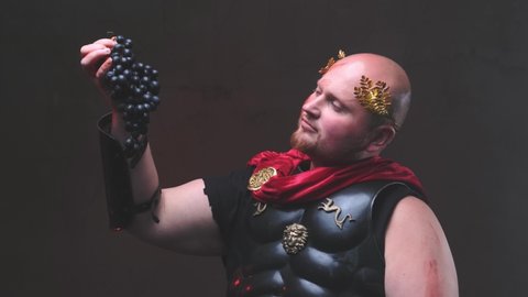 Status and bald joyful legionary with wreath on his had in dark armour with red mantle poses in dark background holding grape and eating it nodding his head.
