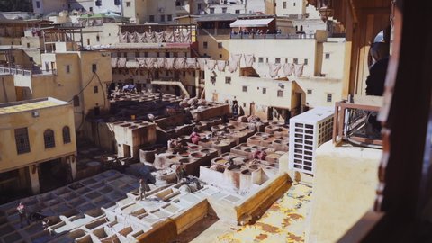 FEZ, MOROCCO - OCTOBER 2020: Elevated Slow Motion Crane Wide Shot Pulling Back Through Balcony Window From People Working In The Chouara Tannery Below To Tourists Watching, Fez, Morocco