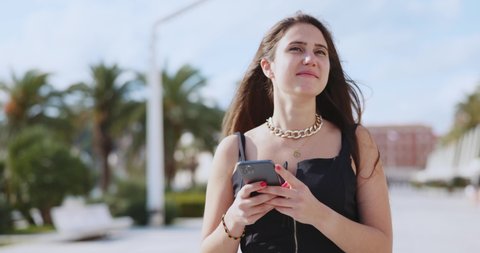 Beautiful young female tourist in elegant dress using mobile phone texting messages browsing social media outdoor. Palm trees. Seaside landscape. Tourism concept.