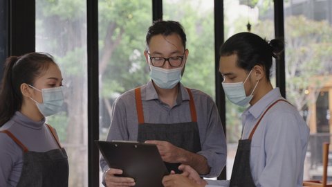 Asian male restaurant manager wearing face mask, explaining and assigning jobs to his colleagues, preparing for reopen after lockdown due to coronavirus pandemic with new normal business concept.
